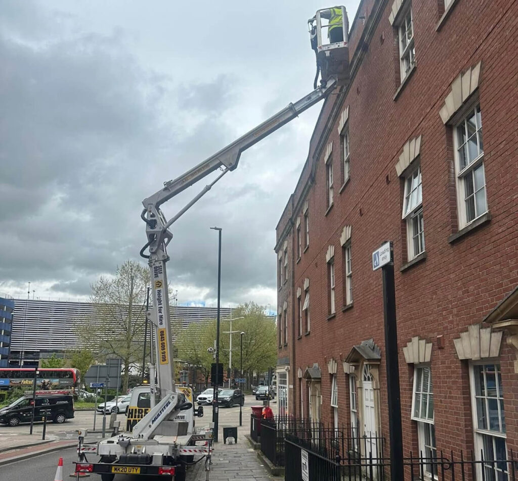 A worker in a lift platform performs maintenance to repair fascia and soffit, as well as the upper windows, on a multi-story brick building along a city street.